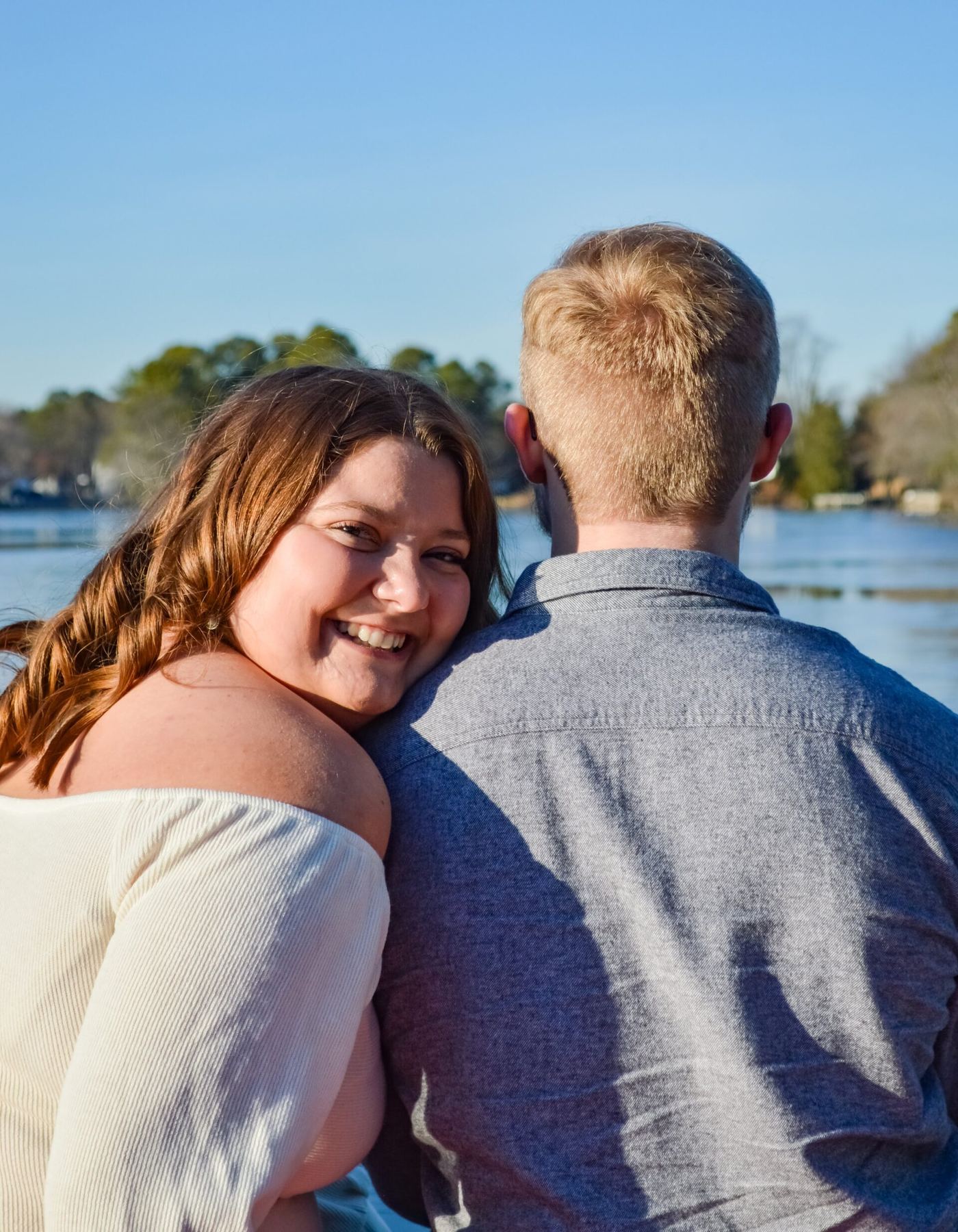 A woman smiling over her partner’s shoulder beside a lake