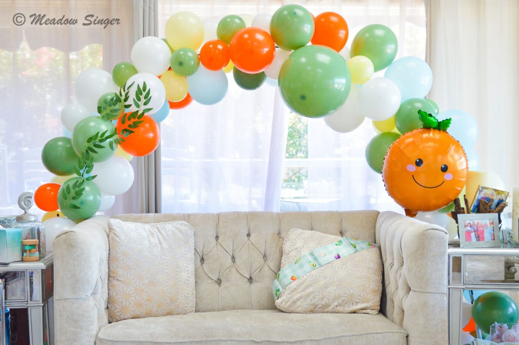 An orange and green balloon arch over a tufted sofa at a baby shower